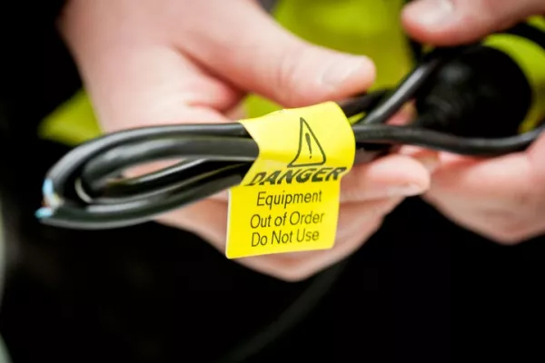 A person holding a damaged black electrical power cable with a yellow "Danger Equipment Out of Order Do Not Use" safety tag attached.