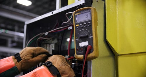 Electrician in protective gloves using a Fluke multimeter to test a commercial electrical panel.
